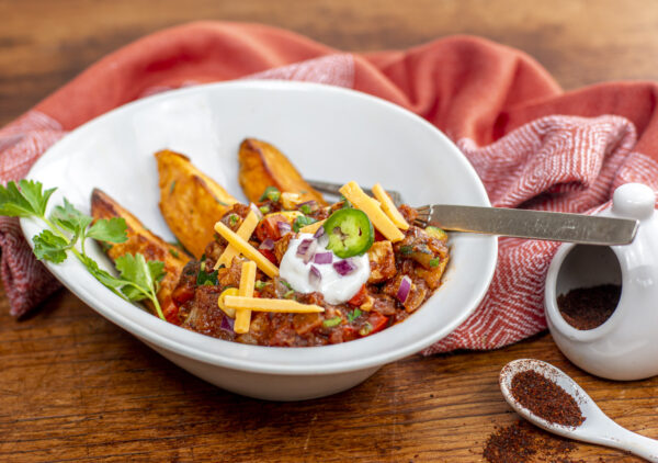 A bowl of Tempeh Chili in a bowl over roasted sweet potato wedges with toppings
